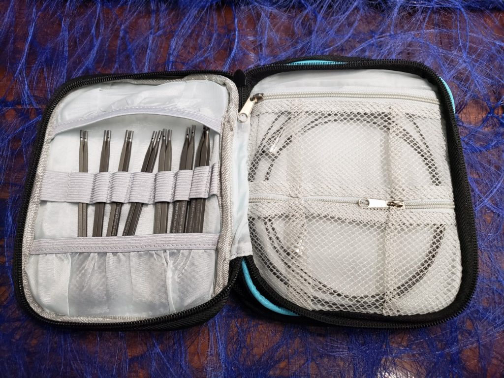 A zippered case lays open on a table, displaying needle tips held in elastic loops and a zippered pocked holding cables and cable stoppers.