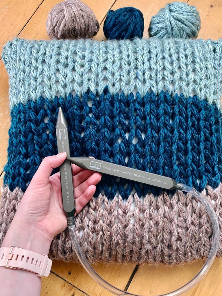 A bulky handknit pillow rests in the background. In the foreground a hand holds a chunky SQUARE fixed circular knitting needle.